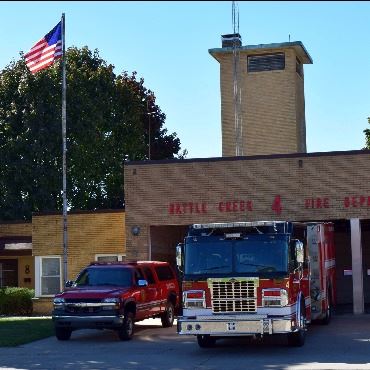 Light brick building in shadow, three fire vehicles parked outside garage. American flag on pole.