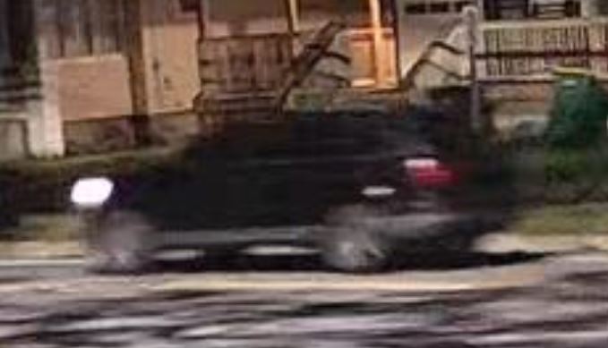 Blurry image of what appears to be a black vehicle on a street in front of a house, at night.
