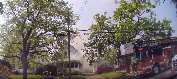 Fire truck parked on neighborhood street next to house showing smoke.